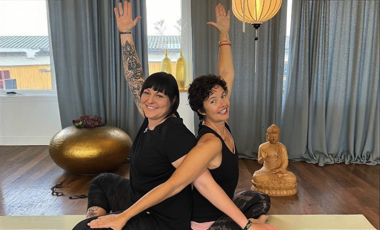 Yoga-Workshop Zwei Frauen sitzen Rücken an Rücken auf Yogamatten in einem hellen Raum und strecken jeweils einen Arm nach oben; im Hintergrund stehen eine Buddha-Figur und ein Gong.